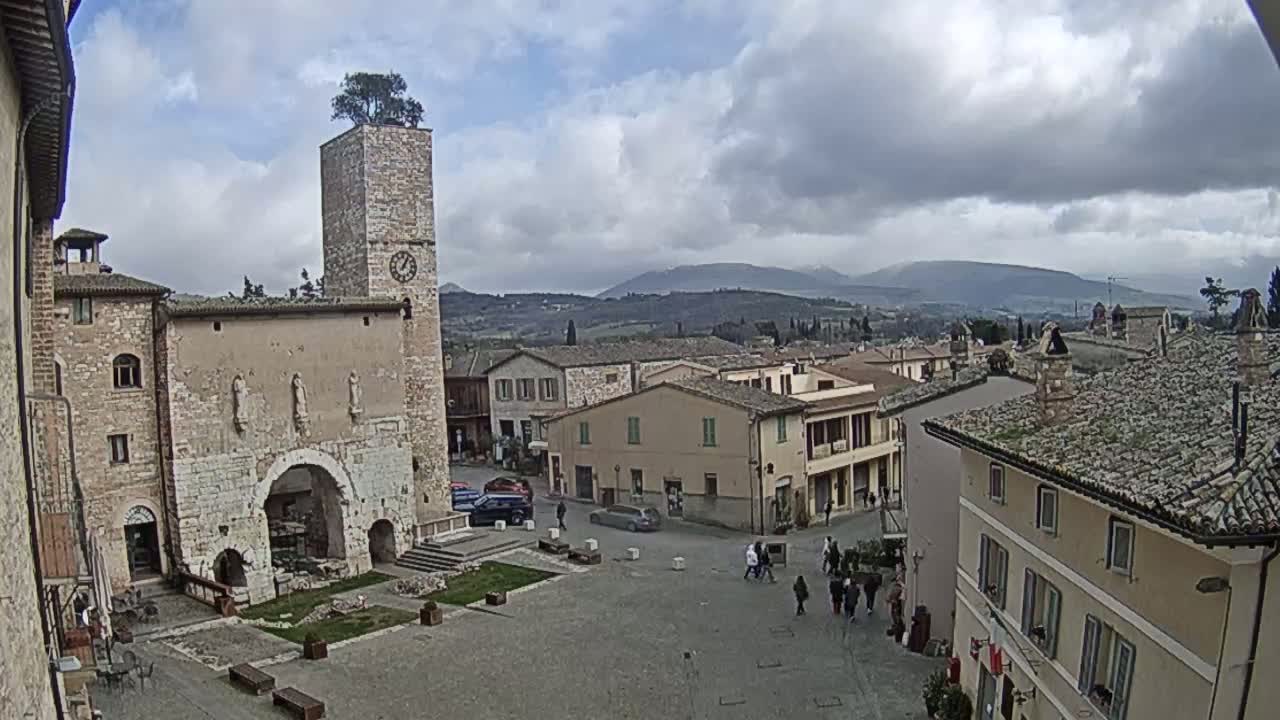 A town square in a hilly, European-style village under a cloudy sky, features a stone tower with a tree on top and several low buildings surrounding the square.