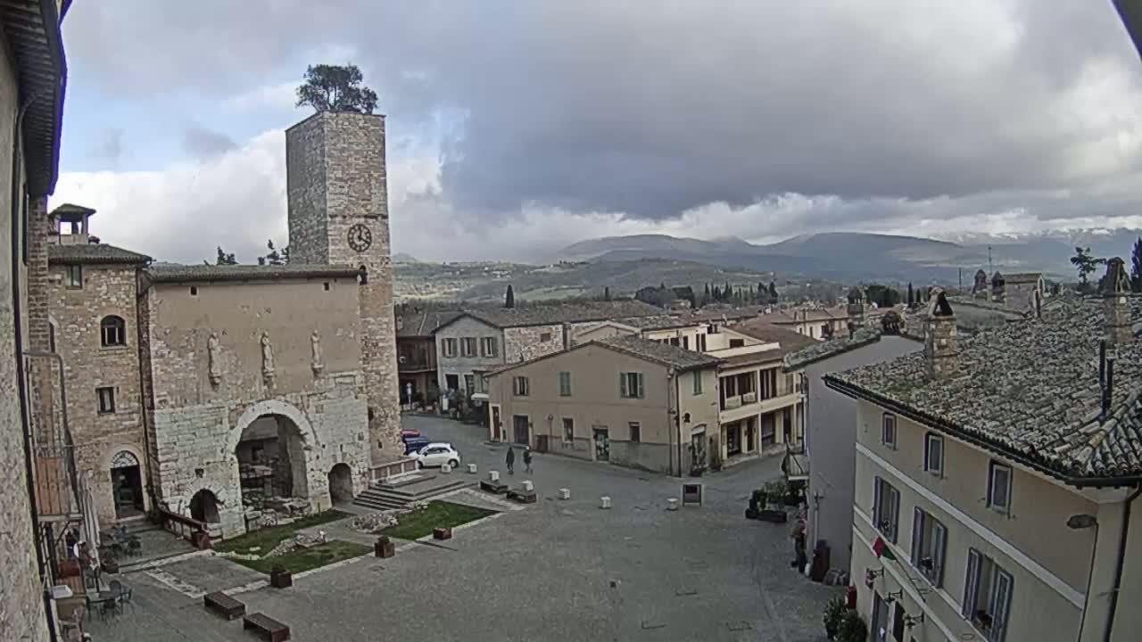 A town square in a historic Italian town, under a cloudy sky, is centered around a large stone building with a clock tower and archway, with other stone buildings and distant hills visible in the background.