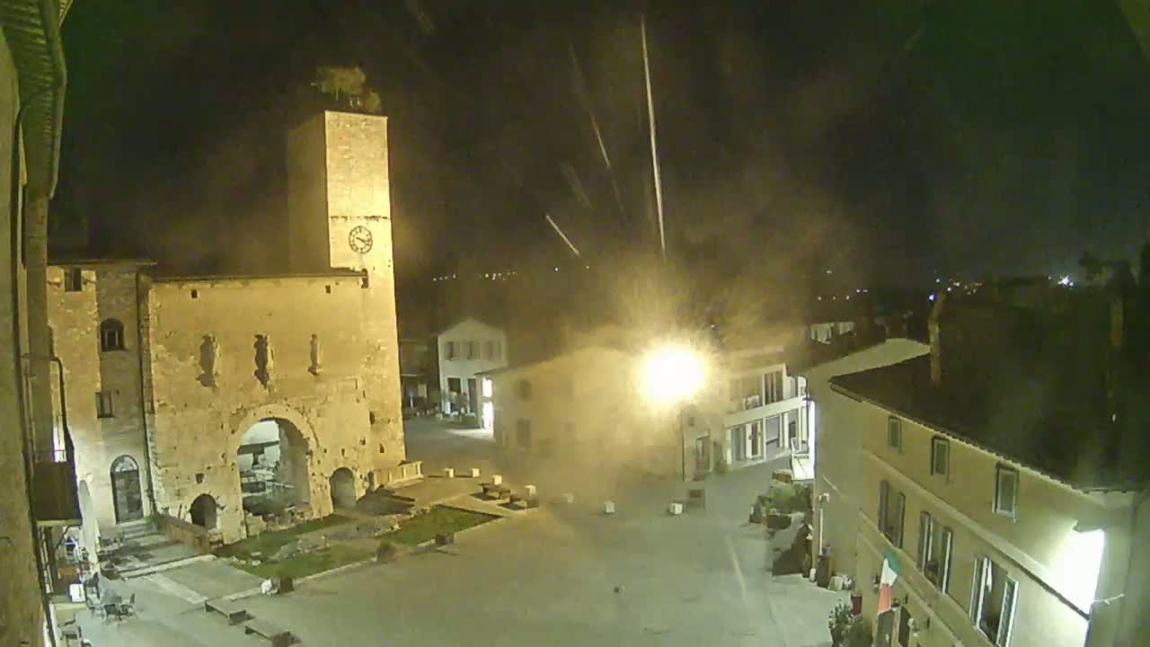 A nighttime view of a town square, featuring an ancient stone archway and tower, under a lightly hazy sky with streaks of light possibly from fireworks.