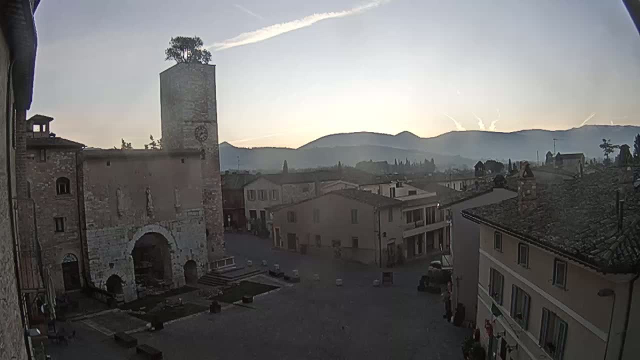 A town square, overlooked by a tall stone tower with a tree on top, sits below hazy mountains under a mostly clear morning sky.