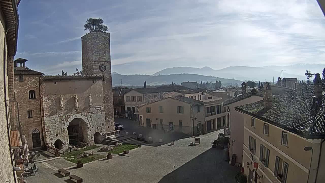 A town square in a historic Italian village, under a mostly clear sky, with mountains visible in the distance.