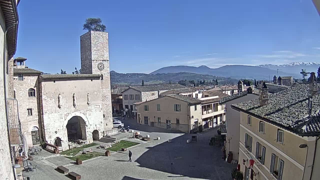 A sunlit town square is surrounded by stone buildings, one featuring a clock tower with a tree on top, under a clear blue sky with distant mountains visible in the background.
