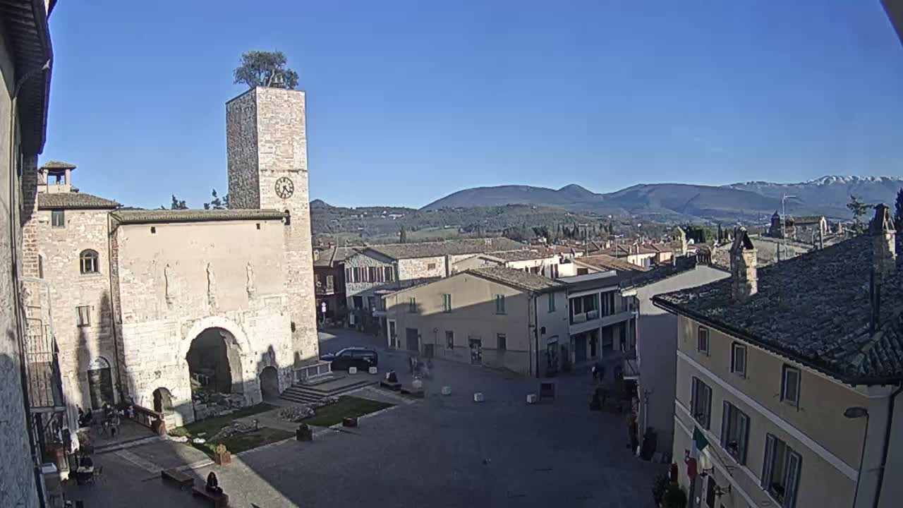 A town square, overlooked by a clock tower with a tree on top, is surrounded by historic buildings under a clear blue sky with distant mountains visible in the background.