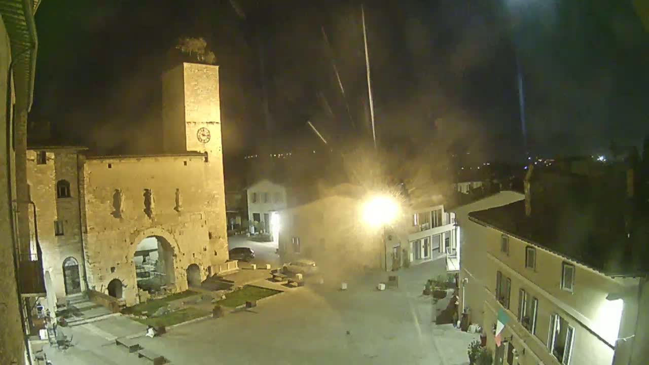 A nighttime view of a town square with a tall clock tower and several buildings, under a hazy sky with streaks of light possibly from fireworks or rain.