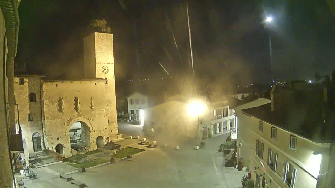 A nighttime view of a town square with stone buildings, including a tall clock tower, under a hazy sky with streaks of light.