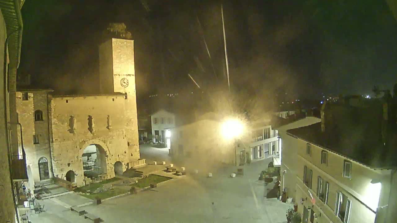 A nighttime view of a town square, illuminated by streetlights, with a tall stone tower and several buildings surrounding it under hazy or foggy conditions.