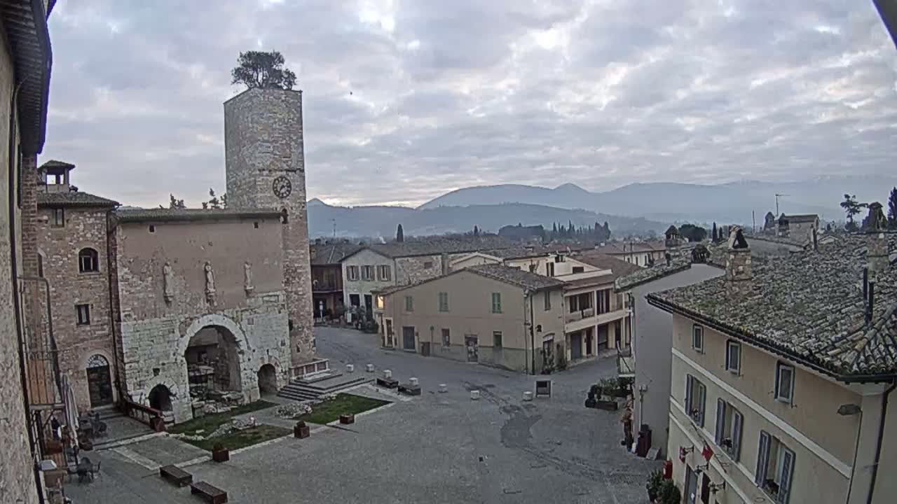 A town square with stone buildings, a clock tower with a tree on top, and a view of distant mountains under a cloudy sky.