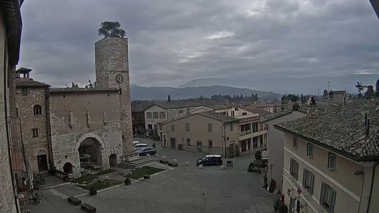 A town square with stone buildings, a clock tower topped with a tree, and distant mountains under a cloudy sky.