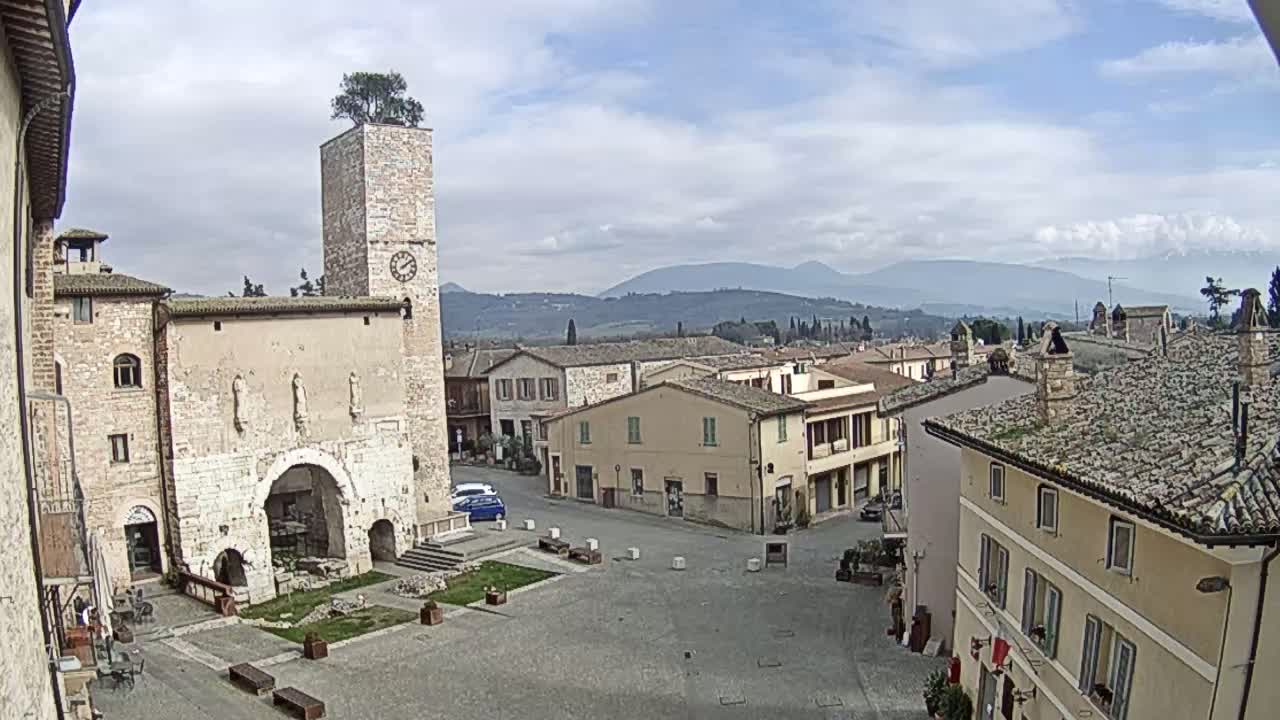 A town square with a stone clock tower, older buildings, and a few cars, under a partly cloudy sky with distant mountains visible in the background.