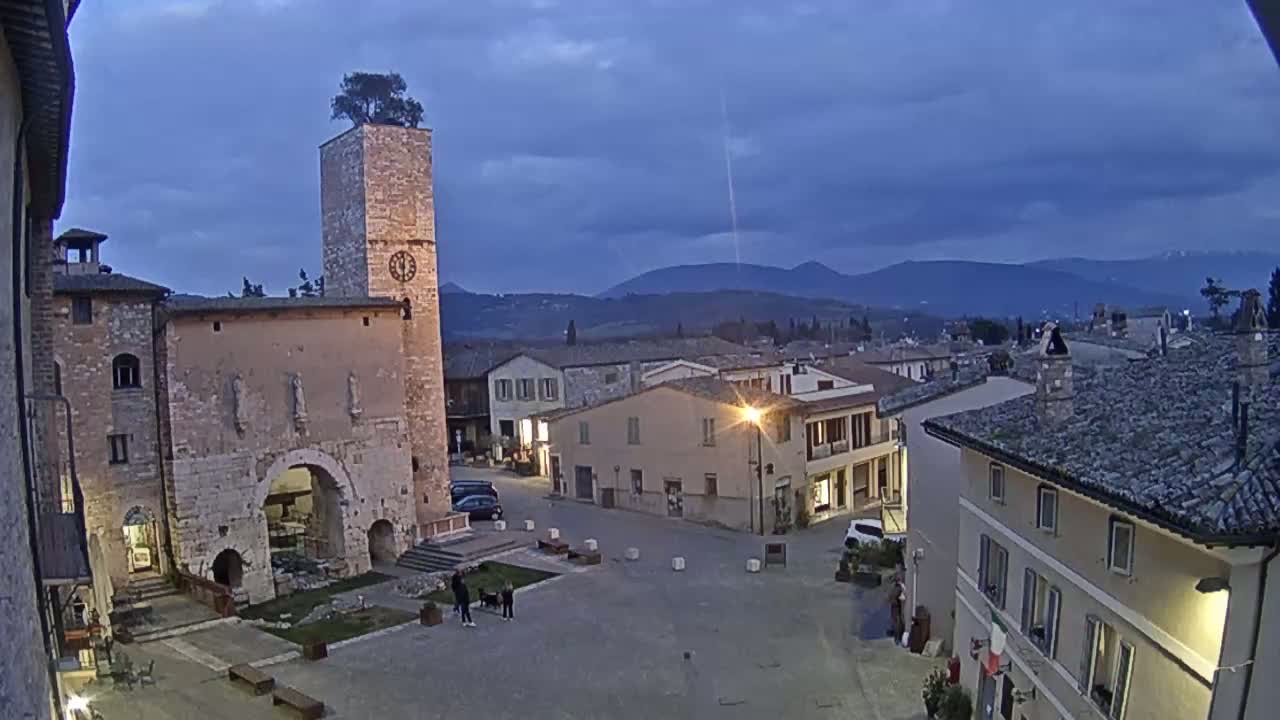 A town square with stone buildings, a clock tower topped with a tree, and distant mountains under a twilight sky.