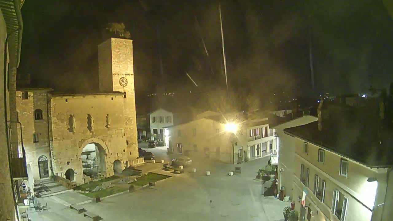 A nighttime view of a town square, featuring an ancient stone archway and clock tower, under a lightly misty or hazy sky with streaks of light possibly from rain or fireworks.