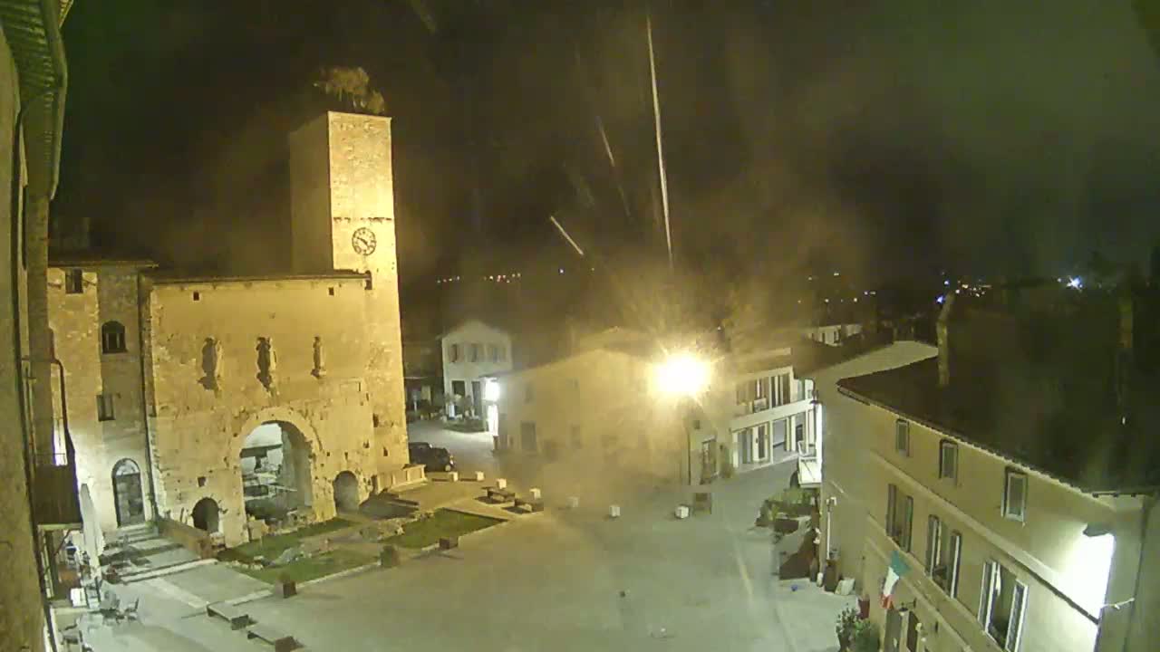 A nighttime, high-angle view of a town square with a tall clock tower and surrounding buildings under a hazy sky.