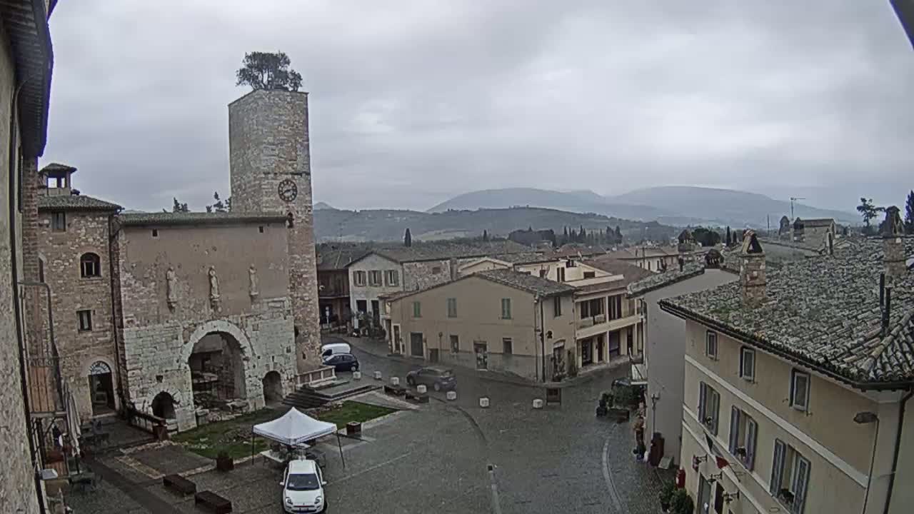 A town square in a historic European village under an overcast sky, featuring a tall stone tower with a tree on top, surrounded by old stone buildings.