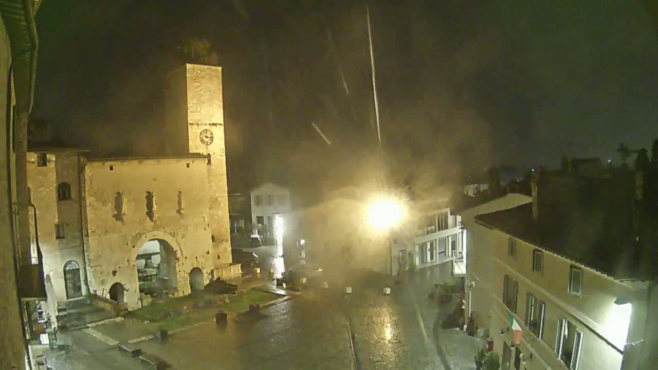 A nighttime view of a town square with a stone tower and buildings under heavy rain.