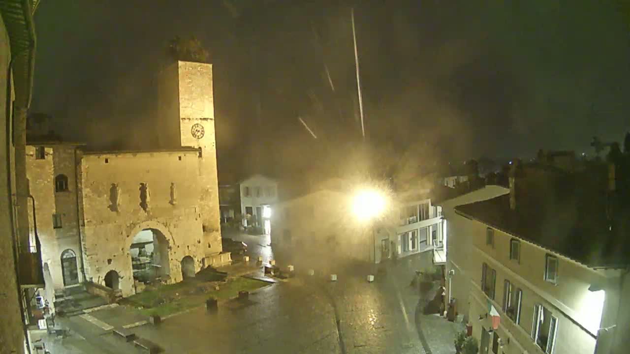 A rain-swept town square at night, illuminated by streetlights, features an ancient stone gate and tower under a dark, stormy sky.
