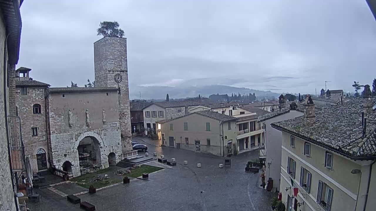 A town square with stone buildings, a clock tower topped with a tree, and a distant view of rolling hills under an overcast sky.