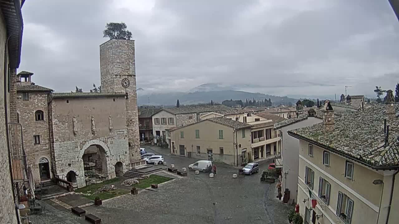 A stone town square, under an overcast sky, is surrounded by old buildings, one with a clock tower topped with a small tree, and overlooks a distant mountain range.