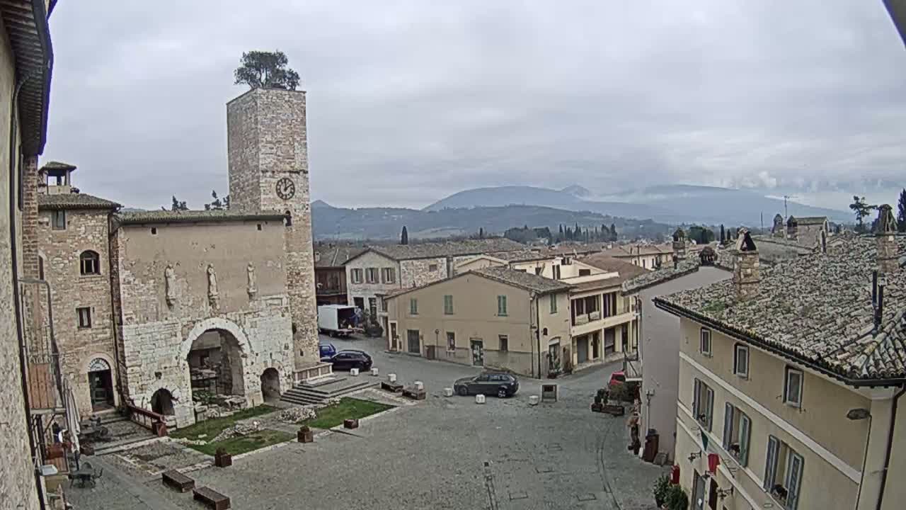 A town square in a European village under an overcast sky, featuring a stone clock tower, several buildings, and a few parked cars.