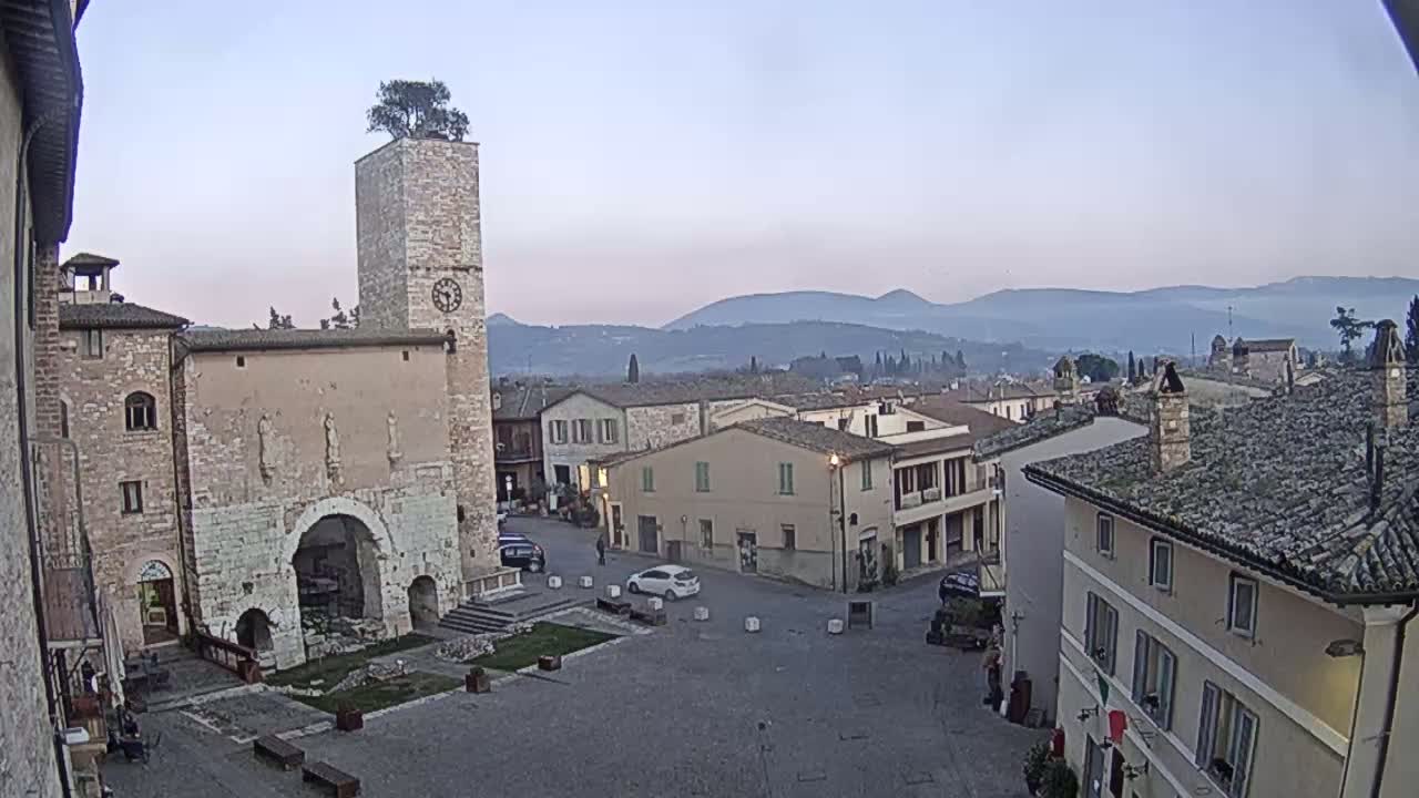 A town square with ancient stone buildings, a clock tower topped with a tree, and a hazy mountain range in the background under a clear, pale sky.