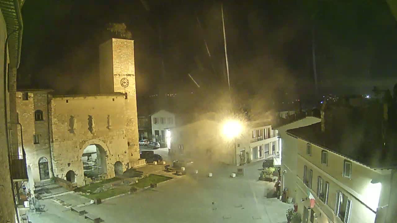 A nighttime view of a town square with old stone buildings, a clock tower, and light rain or snow falling under a dark sky.