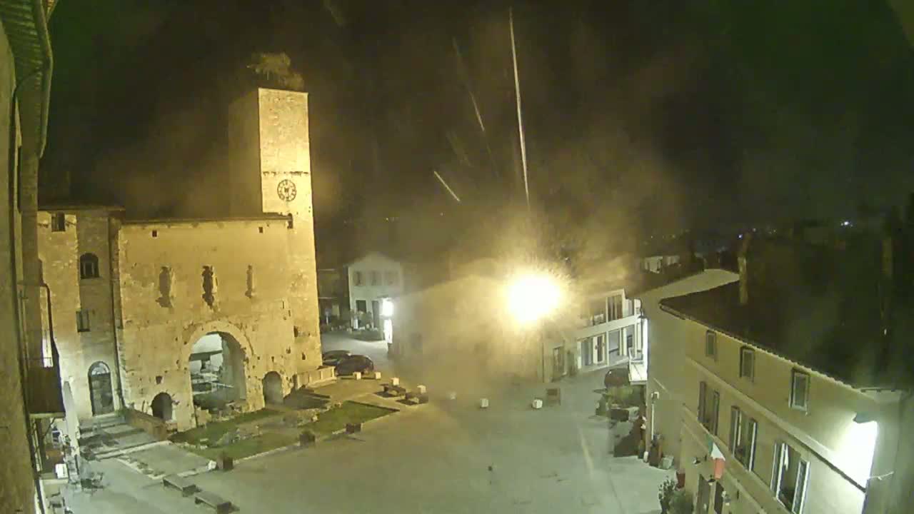 A nighttime view of a town square with an old stone archway and clock tower, illuminated by streetlights, under a hazy sky with what appears to be light rain or snow falling.