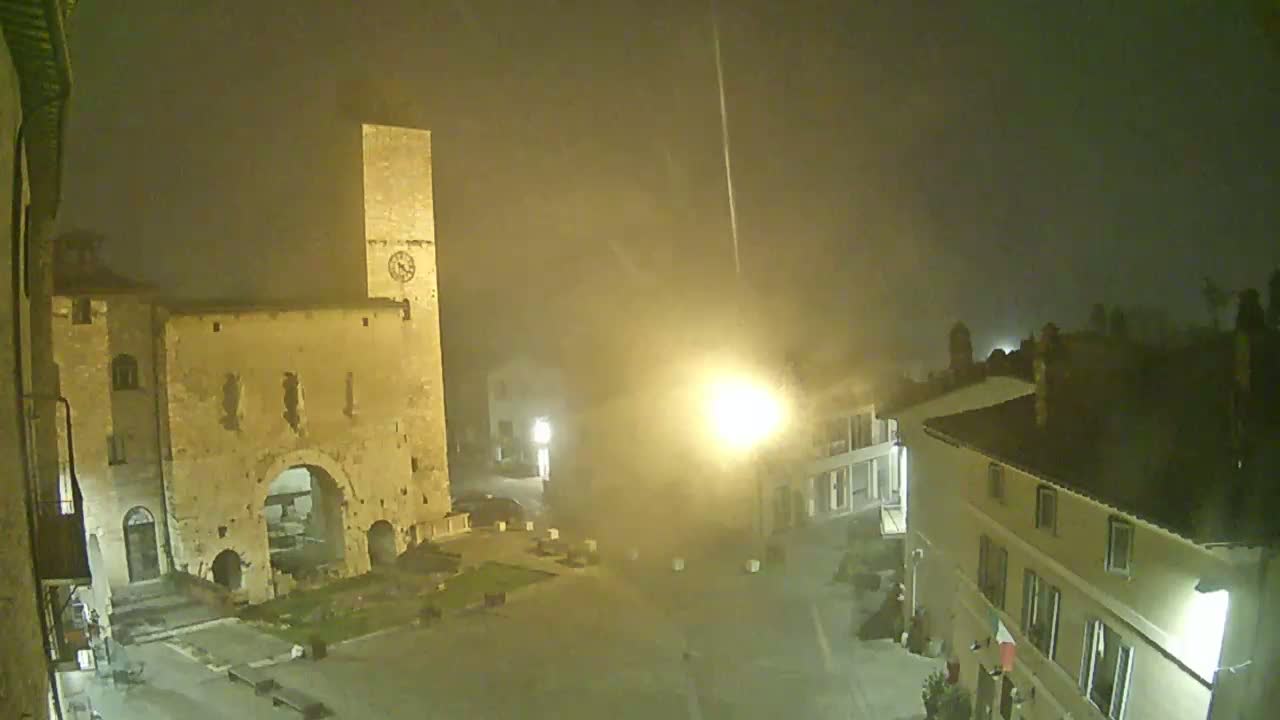 A nighttime, foggy view of a town square, featuring a tall stone tower with a clock, an arched stone structure, and several buildings illuminated by streetlights.