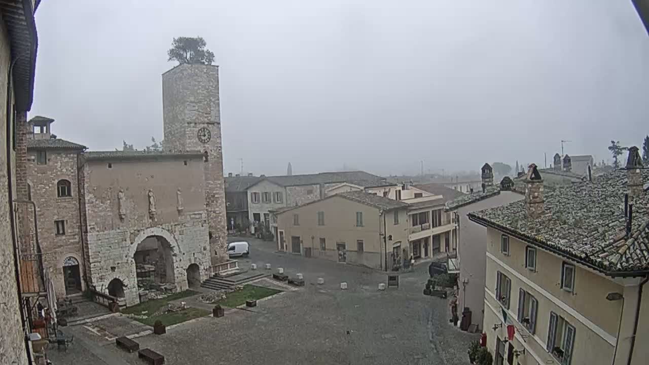 A foggy view of a town square with ancient stone buildings, a clock tower, and a small tree on top of it.