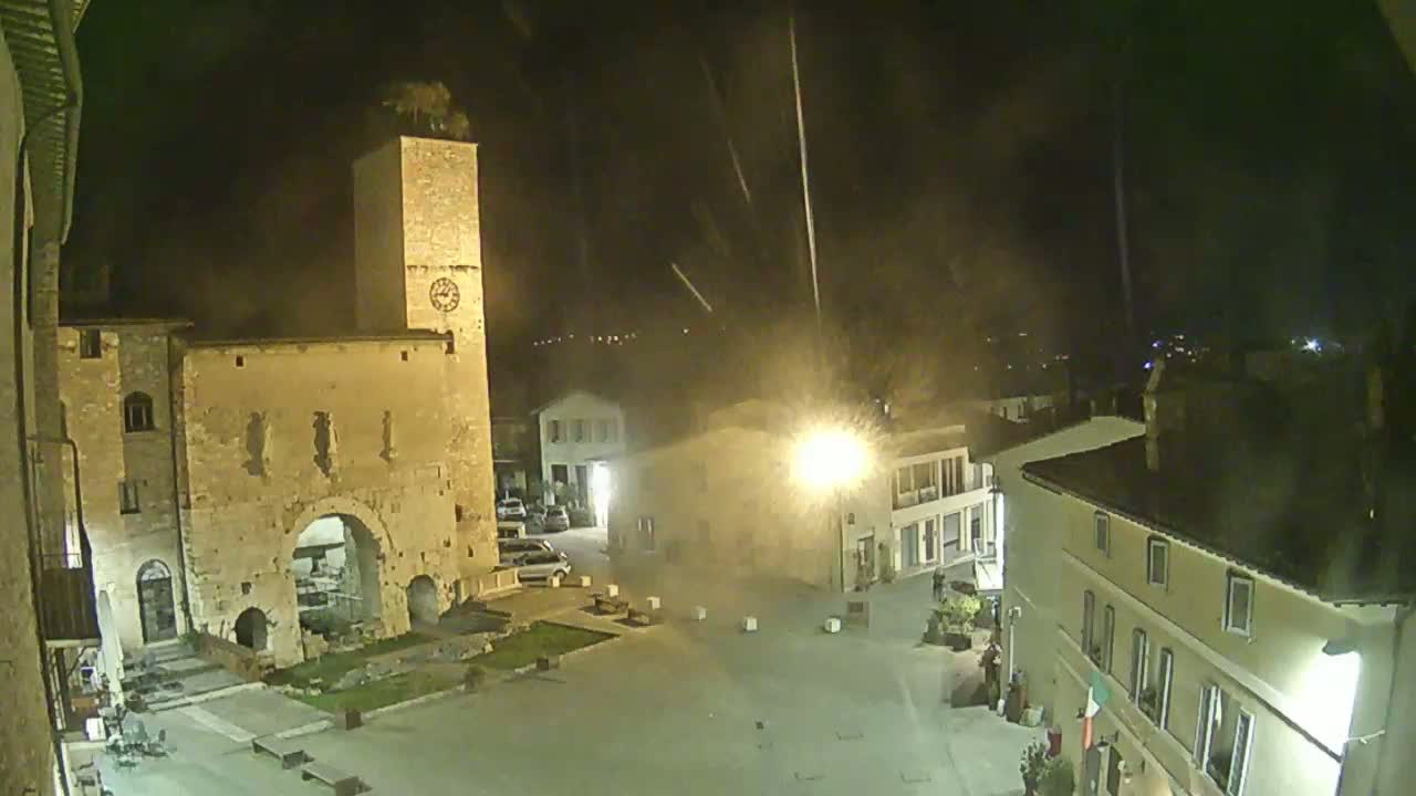 A nighttime view of a town square, illuminated by streetlights, featuring an ancient stone archway and tower, with light rain falling.