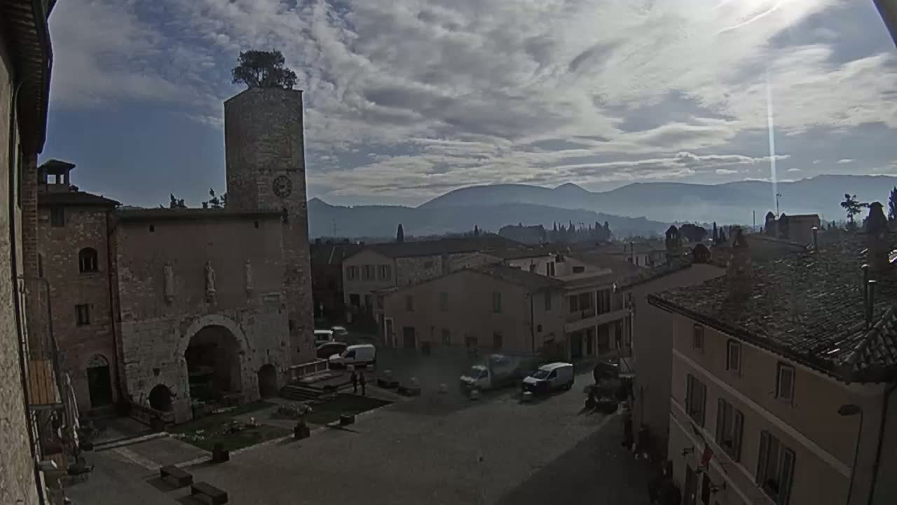 A town square with ancient stone buildings, a clock tower with a tree on top, and a view of hazy mountains in the background under a partly cloudy sky.
