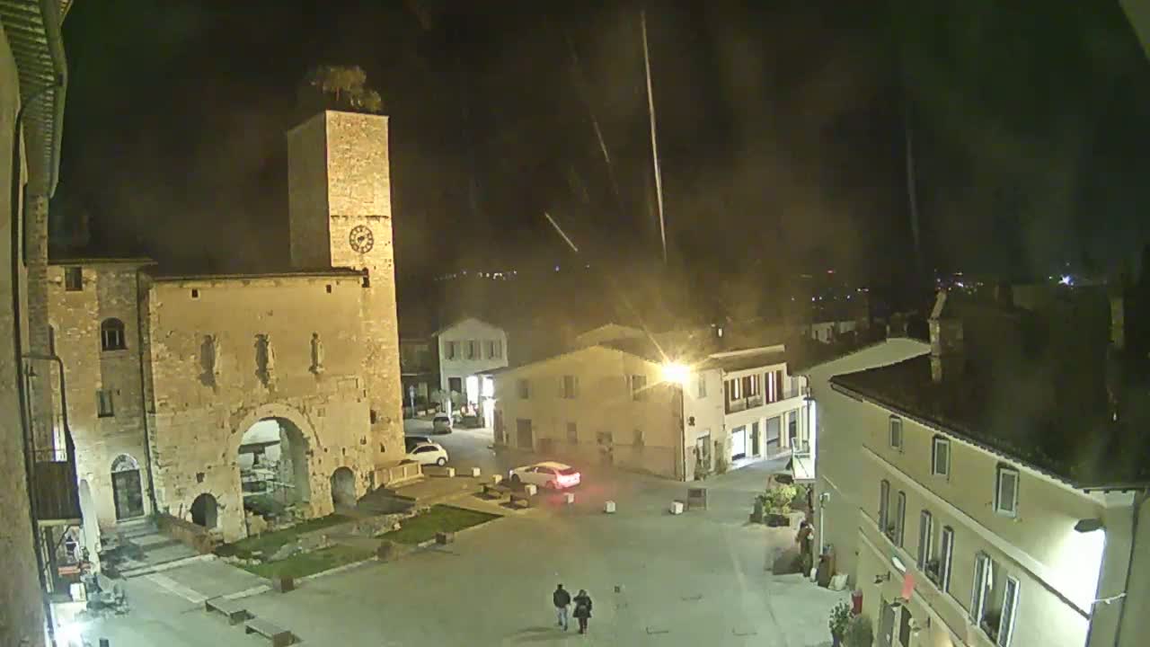 A nighttime view of a town square with a stone tower and several buildings, under a slightly hazy sky with faint streaks of light.