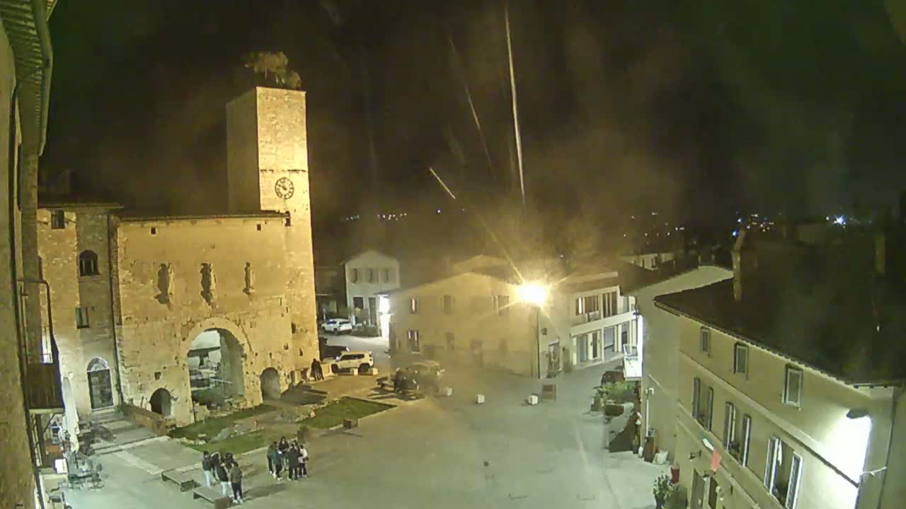 A nighttime view of a town square with a clock tower and several buildings, under slightly hazy conditions.