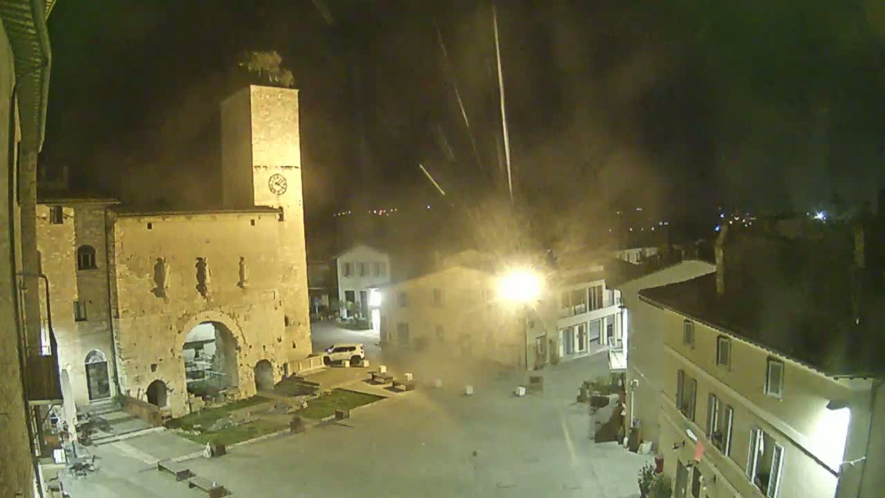 A nighttime view of a town square with stone buildings, a clock tower, and a few cars, under a slightly hazy sky with streaks of what appear to be falling debris.