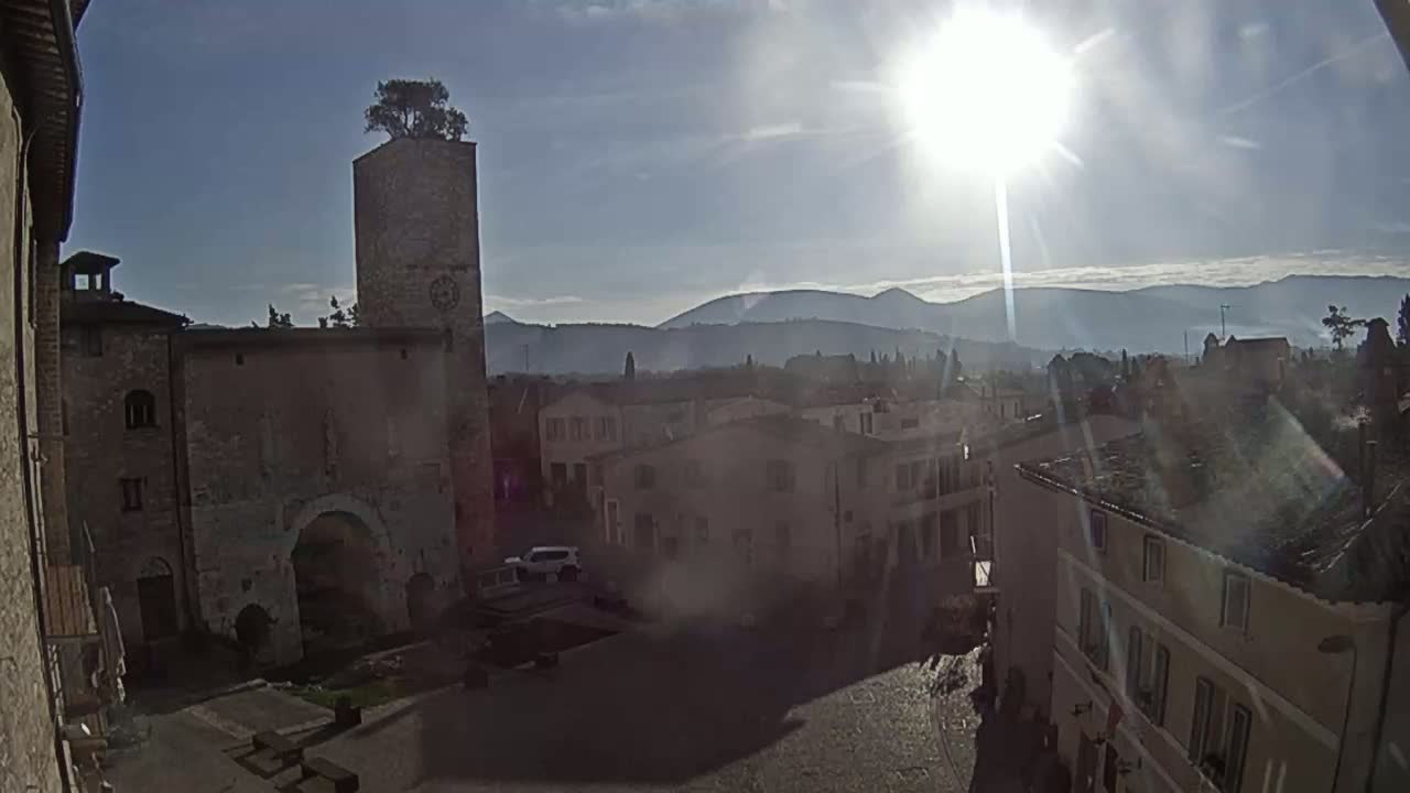 A sunlit view of a town square with ancient stone buildings, a tall tower topped with a tree, and distant mountains under a mostly clear sky.