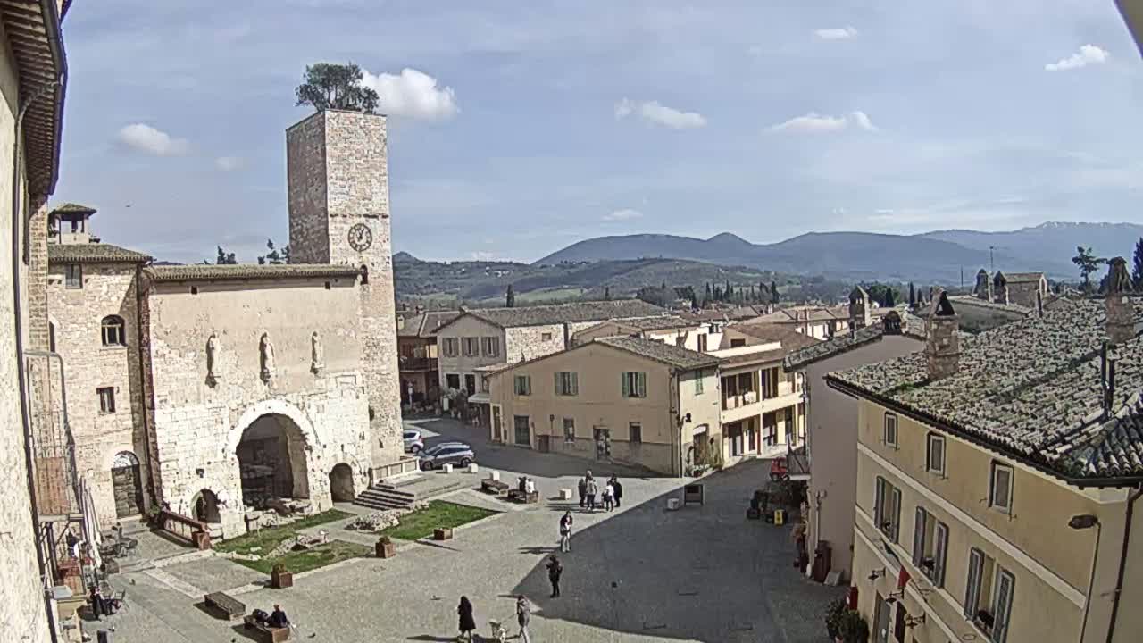 A town square in a sunny, mountainous region features a tall stone tower with a tree on top, surrounded by buildings with terracotta roofs.