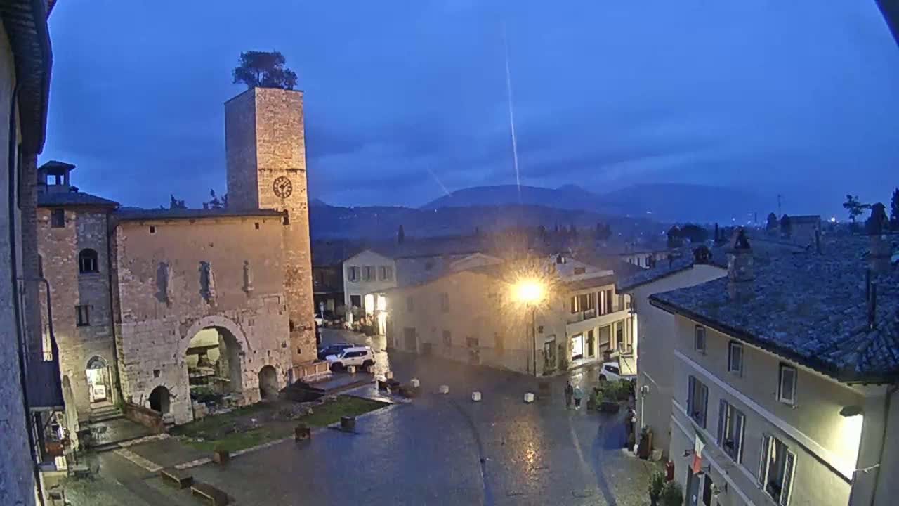 A rain-slicked town square at dusk, centered around an old stone building with a clock tower topped with a tree, is surrounded by other buildings under a twilight sky.