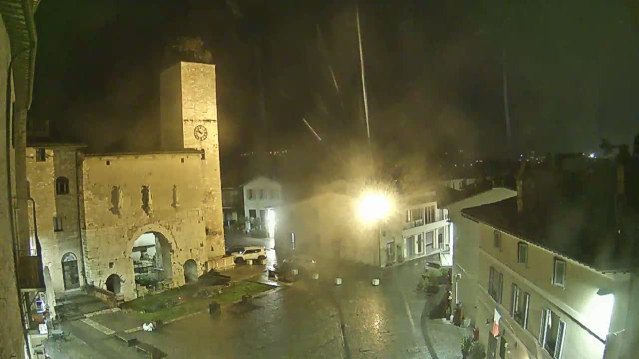 A rain-swept town square at night, illuminated by streetlights, features a medieval stone gate and clock tower, with several buildings surrounding the wet cobblestone plaza.