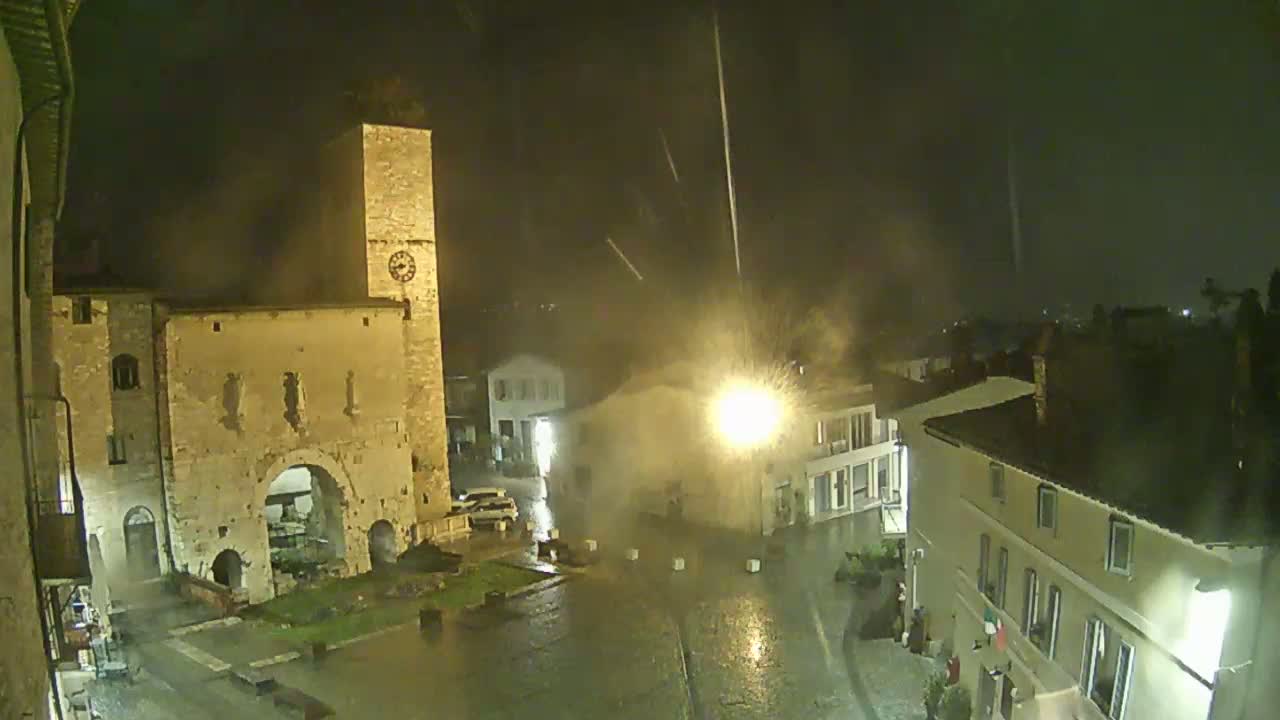 A town square at night during a heavy rain storm is illuminated by streetlights, showcasing a historic stone archway and clock tower.