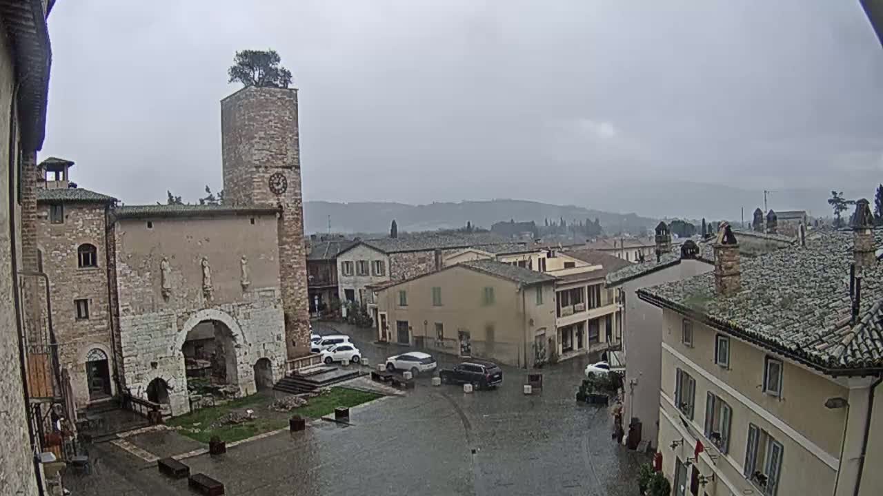 A rain-soaked town square, surrounded by medieval-style buildings, one with a clock tower topped by a small tree, overlooks a distant, hazy landscape.