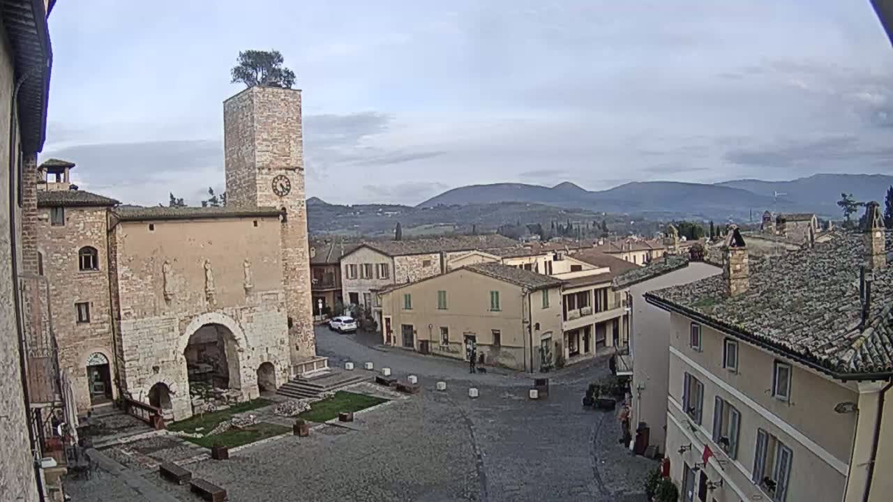 A town square with a stone tower topped with a tree, surrounded by historic buildings, under a cloudy sky and overlooking distant hills.