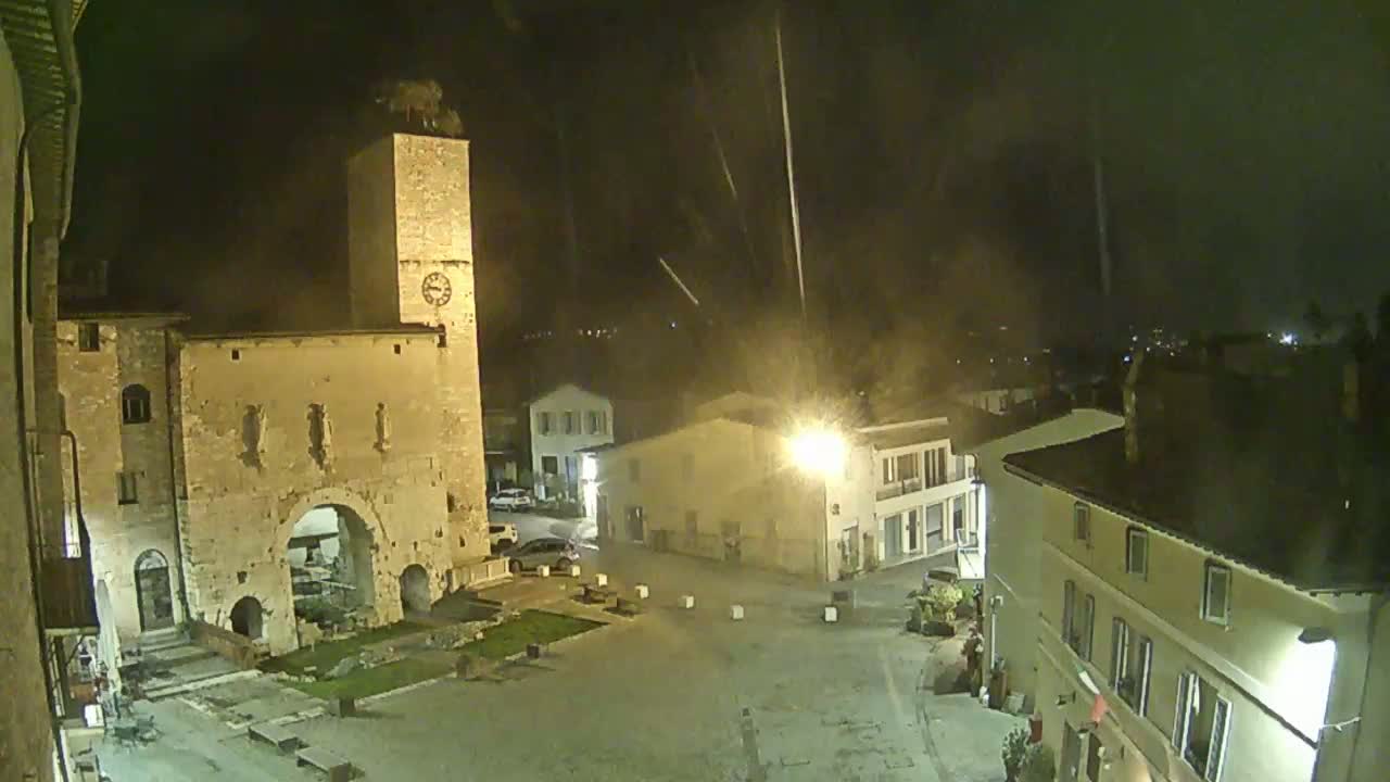 A nighttime view of a town square with ancient stone buildings, a clock tower, and faint streaks in the sky.