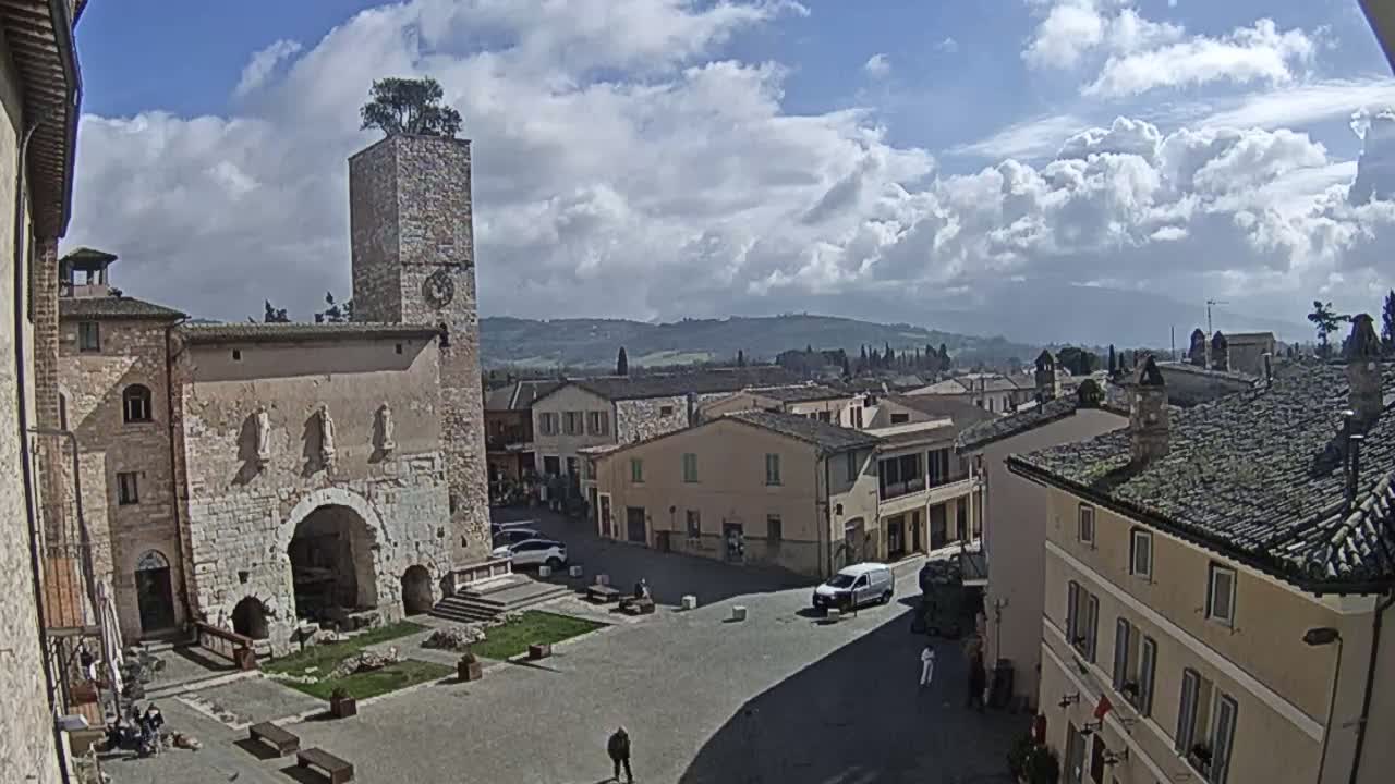 A town square in a historic-looking European town, under a partly cloudy sky, is surrounded by stone buildings, one of which has a tall tower with a tree on top.