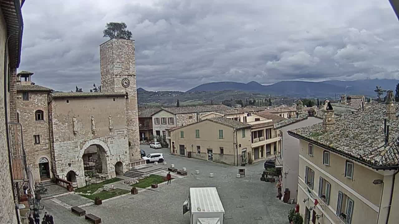 A town square with stone buildings, a clock tower, and a few cars, under an overcast sky with mountains visible in the distance.