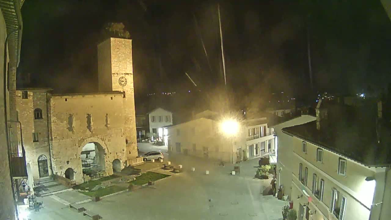 A nighttime view of a town square with old stone buildings, a clock tower, and light rain falling.
