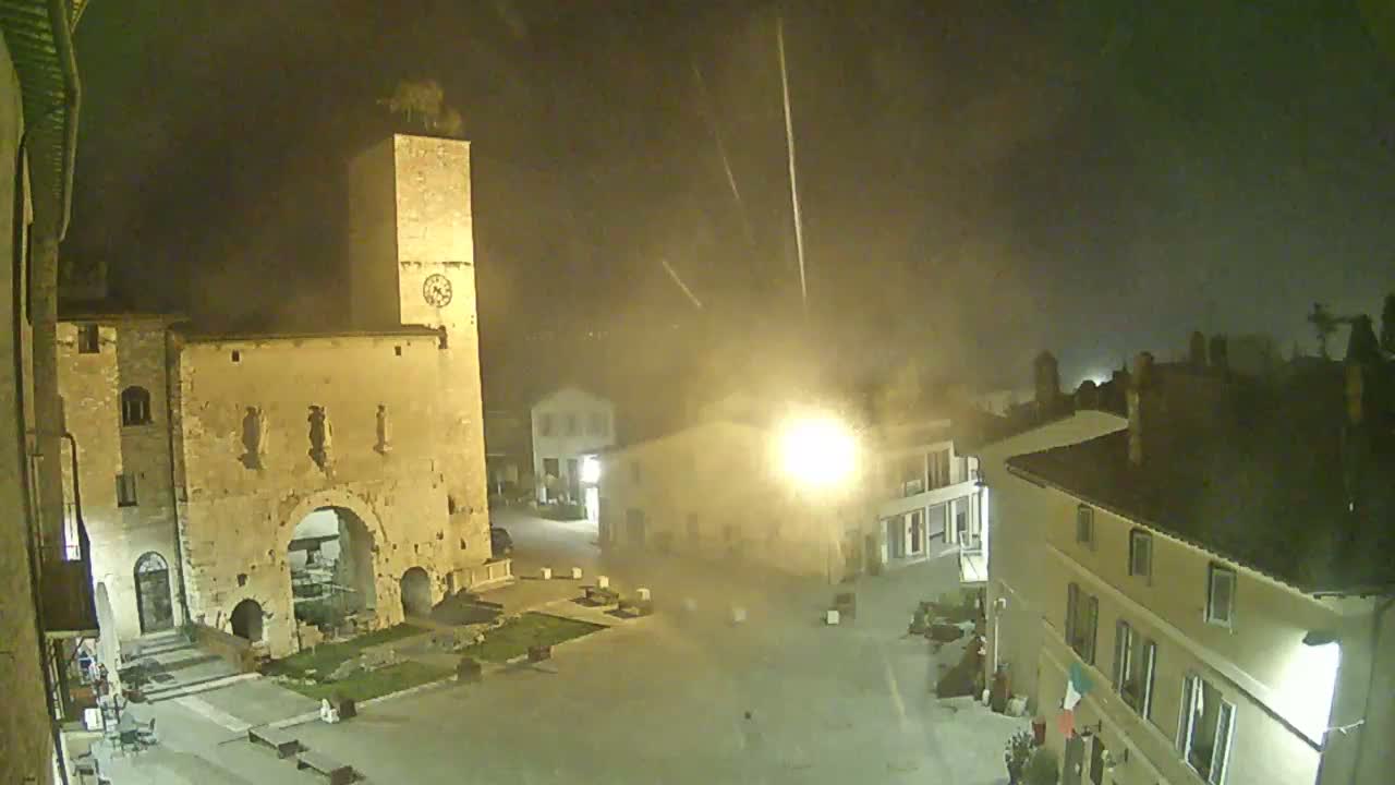 A nighttime view of a town square with ancient stone buildings, including a tall clock tower, under a hazy, possibly rainy sky.