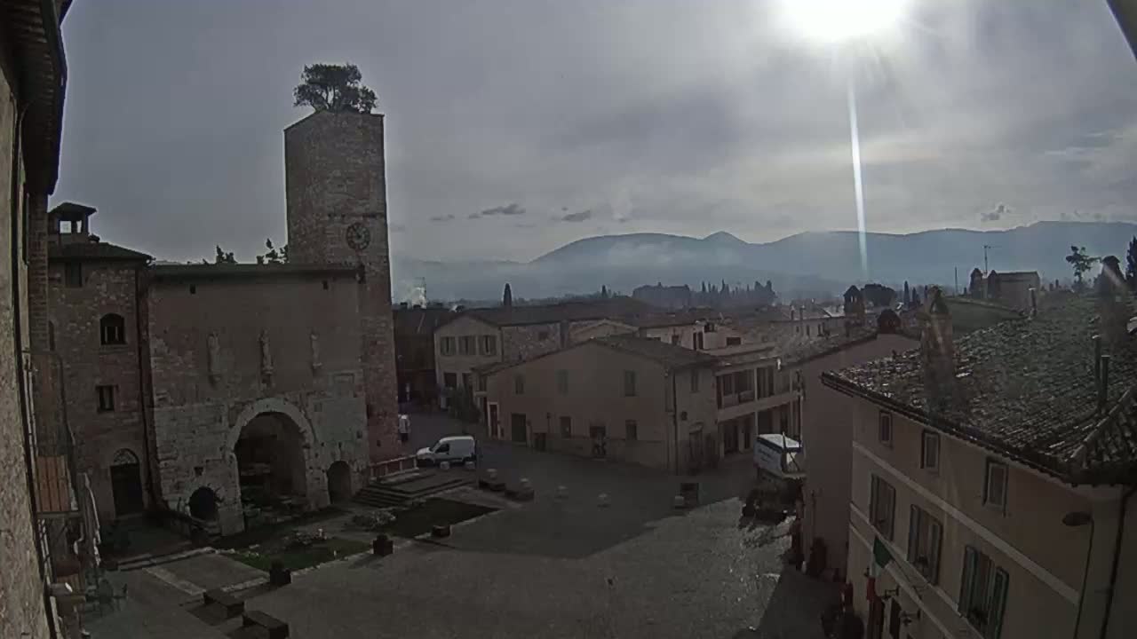 A mostly sunny day reveals a town square with old stone buildings, a clock tower topped with a tree, and distant mountains under a hazy sky.