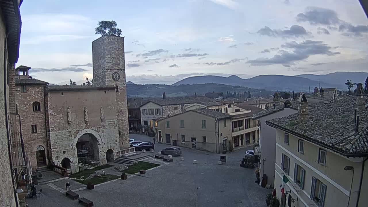 A stone town square with several buildings, including a tall clock tower topped with a tree, overlooks a valley under a mostly cloudy sky.