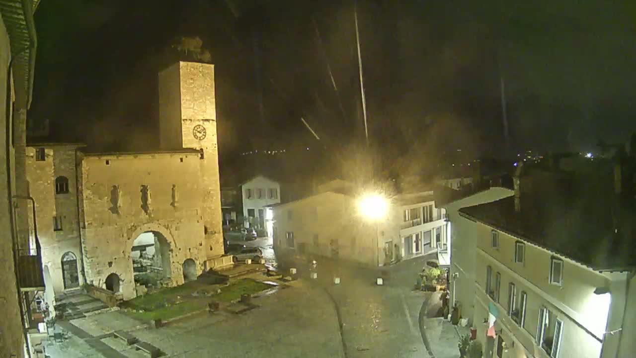 A nighttime view of a town square, illuminated by streetlights, showing an old stone archway, a clock tower, and several buildings under a falling rain or snow.