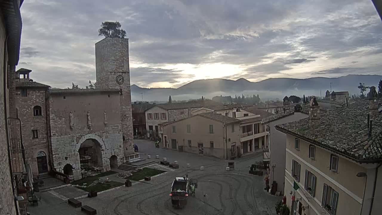 A town square with stone buildings, a tall clock tower with a tree on top, and a street cleaning truck is visible under a partly cloudy, sunrise sky with distant fog-covered mountains.