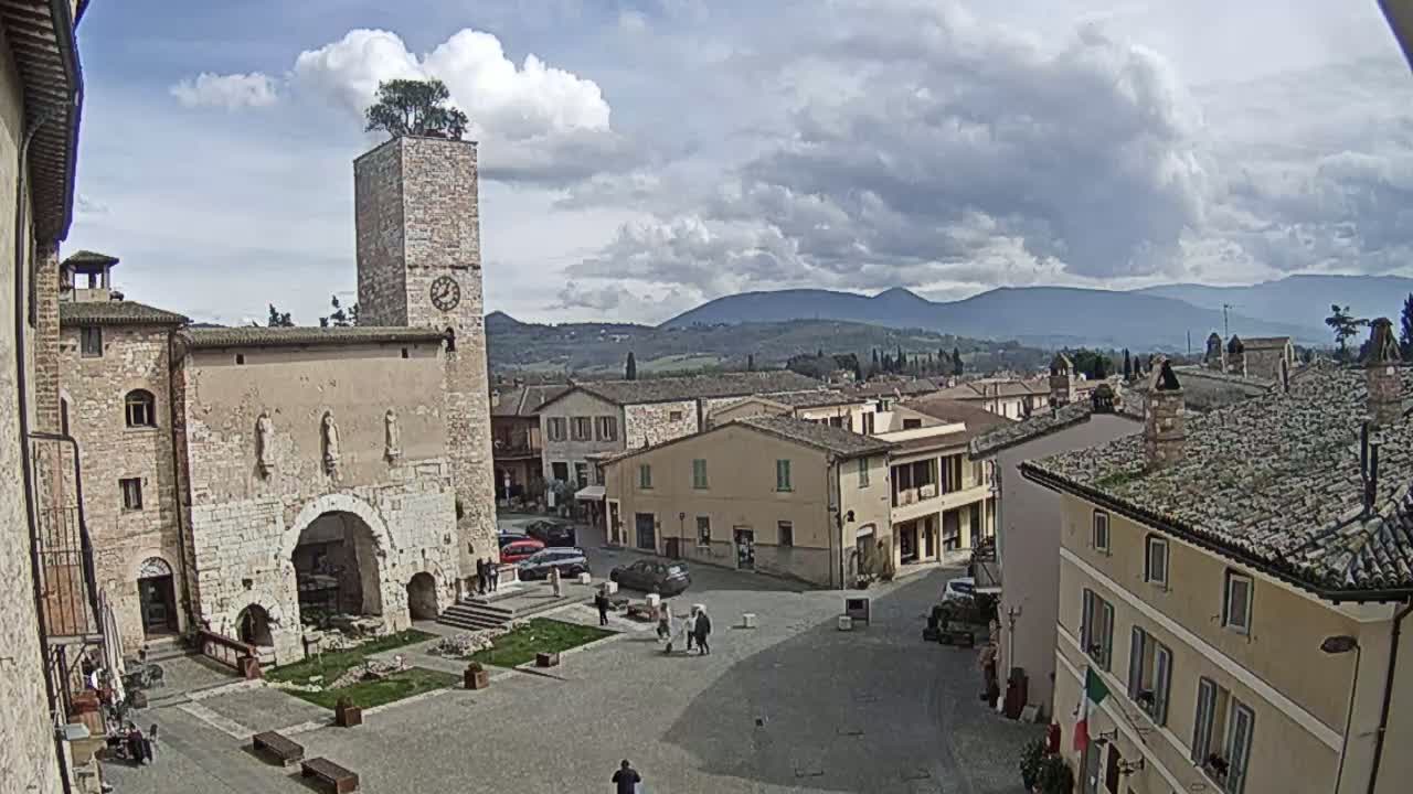 A town square in a hilly, partly cloudy region, centered around an old stone building with a clock tower and a tree on top, features several buildings and a few people.