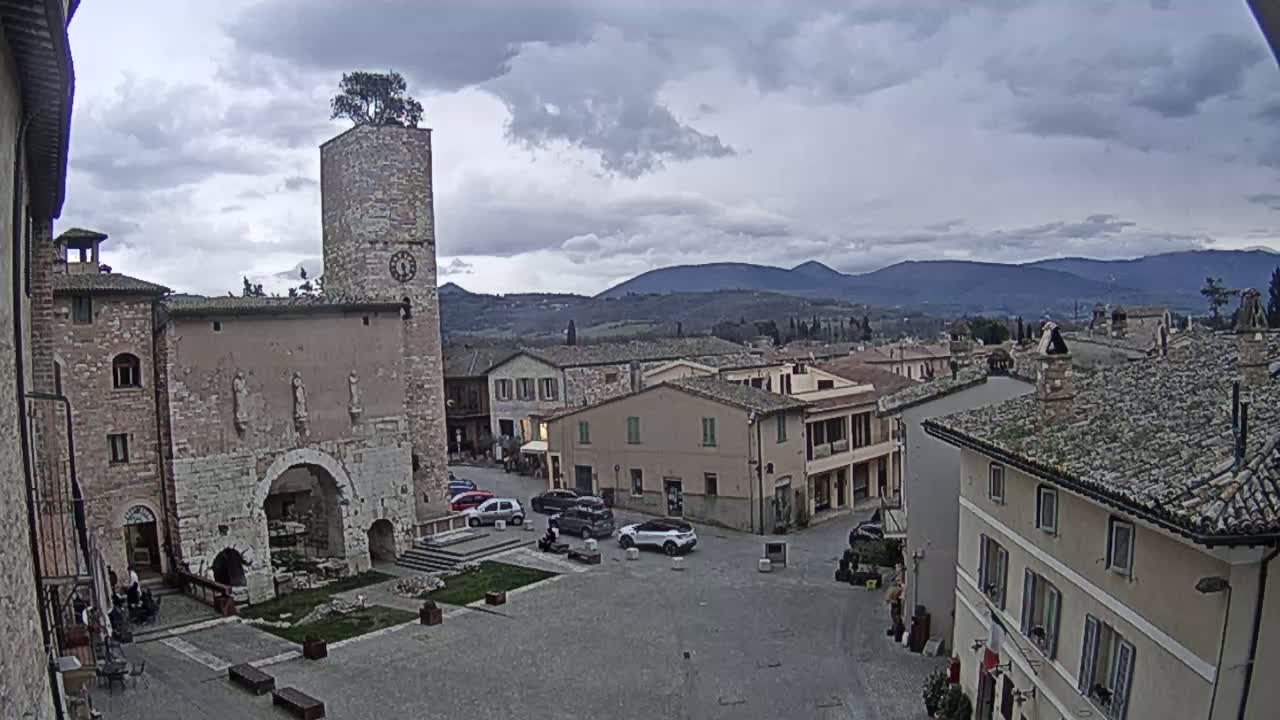 A town square with a brick tower and clock, surrounded by stone buildings, under a cloudy sky and distant mountains.